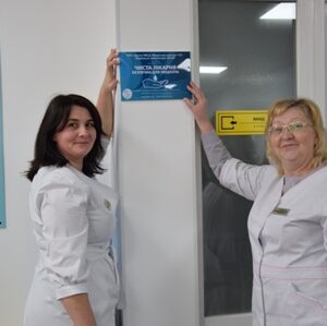 Дві медичні працівниці у білих халатах стоять у приміщенні закладу охорони здоров’я біля дверей та тримають інформаційну табличку на стіні. Позаду видно інформаційні стенди та навігаційні позначки для відвідувачів.