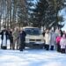 На фото зображена група людей, які стоять на засніженій території на тлі високих дерев. У центрі — світлий мікроавтобус, біля якого зібралися чоловіки та жінки різного віку.