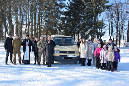 На фото зображена група людей, які стоять на засніженій території на тлі високих дерев. У центрі — світлий мікроавтобус, біля якого зібралися чоловіки та жінки різного віку.