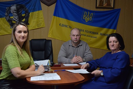 Троє людей сидять за круглим столом у службовому кабінеті та працюють із документами. На столі розкладені папери й канцелярське приладдя. Позаду них на стіні розміщені Державний Прапор України та прапор із гербом і написом про єдність України. Атмосфера робочої наради.