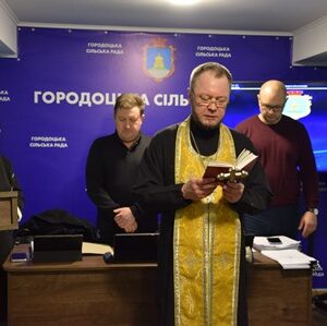 Священник у золотистому облаченні читає молитву з книги під час засідання в приміщенні Городоцької сільської ради. Позаду нього стоять представники керівництва громади та працівники ради. На стіні — синій банер із символікою та написом «Городоцька сільська рада», праворуч розміщений екран.
