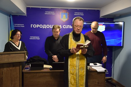 Священник у золотистому облаченні читає молитву з книги під час засідання в приміщенні Городоцької сільської ради. Позаду нього стоять представники керівництва громади та працівники ради. На стіні — синій банер із символікою та написом «Городоцька сільська рада», праворуч розміщений екран.