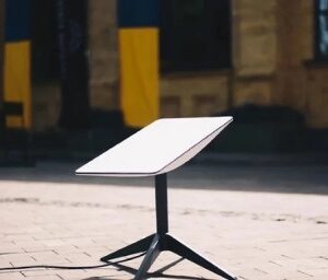 На фото — інформаційна табличка на металевій стійці, встановлена на вимощеній площі. На задньому плані видно будівлю з колонами та вертикальні елементи у синьо-жовтих кольорах.