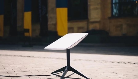 На фото — інформаційна табличка на металевій стійці, встановлена на вимощеній площі. На задньому плані видно будівлю з колонами та вертикальні елементи у синьо-жовтих кольорах.