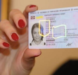 Жінка з червоним манікюром тримає в руці посвідку на тимчасове проживання в Україні. На пластиковій картці видно фотографію власниці, її персональні дані, номер документа, голографічний захисний елемент та зазначений термін дії. Фон розмитий, теплих відтінків.