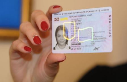 Жінка з червоним манікюром тримає в руці посвідку на тимчасове проживання в Україні. На пластиковій картці видно фотографію власниці, її персональні дані, номер документа, голографічний захисний елемент та зазначений термін дії. Фон розмитий, теплих відтінків.