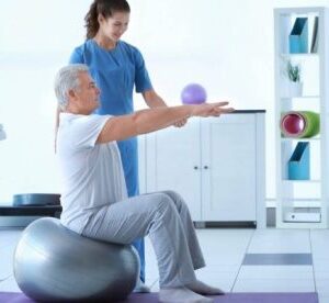 Літній чоловік сидить на фітболі та виконує вправу з витягнутими вперед руками під наглядом реабілітолога в світлому кабінеті лікувальної фізкультури.