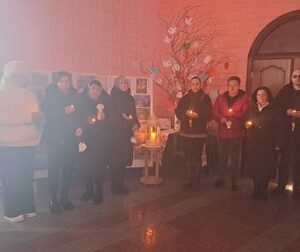 Група людей стоїть у приміщенні з рожевими стінами та тримає запалені свічки в руках. У центрі — невеликий столик із ліхтарем зі свічкою всередині, поруч стенд із фотографіями.