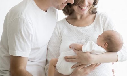 Молода сім’я тримає на руках новонароджену дитину та усміхається.