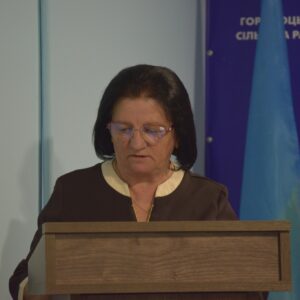 Жінка середнього віку з темним волоссям і в окулярах стоїть за дерев’яною трибуною та читає з аркуша або нотаток. На ній темний одяг із світлою вставкою біля коміра. Позаду видно синій банер із написом українською мовою та прапор. Атмосфера нагадує офіційний виступ або засідання.