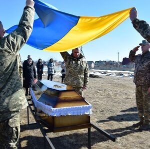 Військовослужбовці тримають Державний Прапор України над труною загиблого Захисника під час церемонії прощання на відкритій місцевості.