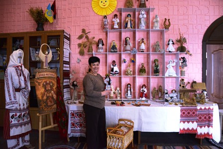 Жінка у вишиванці стоїть біля експозиції ляльок-мотанок у Будинку культури: на столі та полицях представлені різноманітні традиційні ляльки, поруч — декоративні елементи українського інтер’єру та інформаційний стенд.