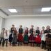 «Група дітей і дорослих у традиційному українському одязі співають у приміщенні, утворивши півколо; деякі учасники тримають музичні інструменти»
