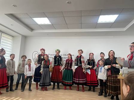 «Група дітей і дорослих у традиційному українському одязі співають у приміщенні, утворивши півколо; деякі учасники тримають музичні інструменти»