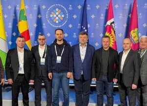 Група чоловіків і жінка стоять разом для спільного фото на тлі синього банера з логотипом та прапорами різних регіонів; учасники заходу у діловому одязі з бейджами.