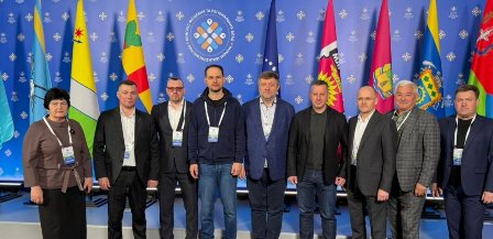 Група чоловіків і жінка стоять разом для спільного фото на тлі синього банера з логотипом та прапорами різних регіонів; учасники заходу у діловому одязі з бейджами.