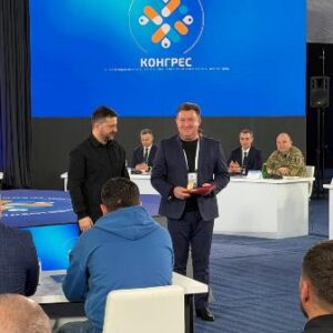 На фото зображено момент офіційного заходу: у конференц-залі на сцені двоє чоловіків стоять поруч, один із них вручає іншому нагороду або відзнаку.