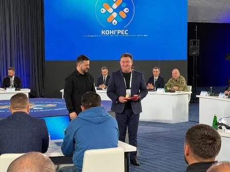 На фото зображено момент офіційного заходу: у конференц-залі на сцені двоє чоловіків стоять поруч, один із них вручає іншому нагороду або відзнаку.