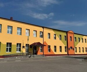 Сучасна двоповерхова будівля навчального закладу, пофарбована у жовто-оранжеві кольори, з великими вікнами та центральним входом із навісом. Перед будівлею — просторе подвір’я з асфальтованим покриттям, поруч видно дерева та стовп освітлення.