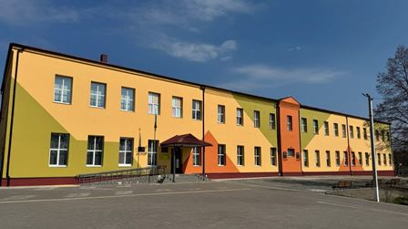 Сучасна двоповерхова будівля навчального закладу, пофарбована у жовто-оранжеві кольори, з великими вікнами та центральним входом із навісом. Перед будівлею — просторе подвір’я з асфальтованим покриттям, поруч видно дерева та стовп освітлення.