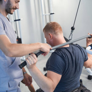 Чоловік під час реабілітаційного тренування виконує вправу на тренажері з тягою за голову, сидячи обличчям від обладнання. Поруч стоїть реабілітолог у медичній формі, який контролює виконання вправи