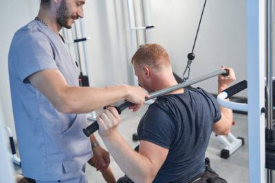 Чоловік під час реабілітаційного тренування виконує вправу на тренажері з тягою за голову, сидячи обличчям від обладнання. Поруч стоїть реабілітолог у медичній формі, який контролює виконання вправи