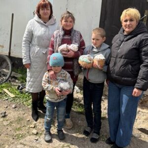 На фото четверо жінок і двоє дітей стоять біля будинку на подвір’ї. У руках вони тримають великодні паски, загорнуті в пакети. Люди одягнені в верхній одяг, поруч видно велосипед і господарські речі. Сцена передає момент передачі святкових гостинців.