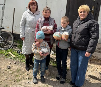 На фото четверо жінок і двоє дітей стоять біля будинку на подвір’ї. У руках вони тримають великодні паски, загорнуті в пакети. Люди одягнені в верхній одяг, поруч видно велосипед і господарські речі. Сцена передає момент передачі святкових гостинців.