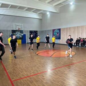 Гравці під час баскетбольного матчу у спортивній залі, на лаві сидять глядачі.