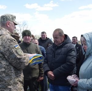 Військовослужбовець у камуфляжі тримає folded державний прапор і спілкується з рідними під час церемонії прощання на відкритому повітрі. Навколо стоять люди в скорботі.