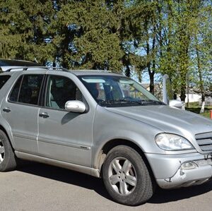 Сріблястий позашляховик Mercedes-Benz ML-класу припаркований на узбіччі дороги. Автомобіль має відкритий багажник, на даху встановлені рейлінги. Поруч видно зелені дерева та огорожу, на задньому плані — приватні будівлі.