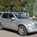 Сріблястий позашляховик Mercedes-Benz ML-класу припаркований на узбіччі дороги. Автомобіль має відкритий багажник, на даху встановлені рейлінги. Поруч видно зелені дерева та огорожу, на задньому плані — приватні будівлі.