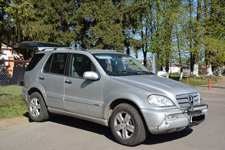 Сріблястий позашляховик Mercedes-Benz ML-класу припаркований на узбіччі дороги. Автомобіль має відкритий багажник, на даху встановлені рейлінги. Поруч видно зелені дерева та огорожу, на задньому плані — приватні будівлі.