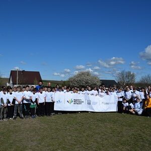 Групове фото учасників спортивного заходу на відкритому стадіоні: кілька десятків дітей і дорослих у білих футболках стоять разом на траві, тримаючи великий банер із логотипами проєкту.