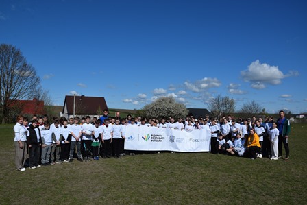 Групове фото учасників спортивного заходу на відкритому стадіоні: кілька десятків дітей і дорослих у білих футболках стоять разом на траві, тримаючи великий банер із логотипами проєкту.