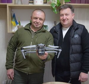 Двоє чоловіків у приміщенні стоять поруч і тримають у руках квадрокоптер; на тлі — полиці з декором та прапор України.