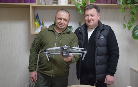 Двоє чоловіків у приміщенні стоять поруч і тримають у руках квадрокоптер; на тлі — полиці з декором та прапор України.