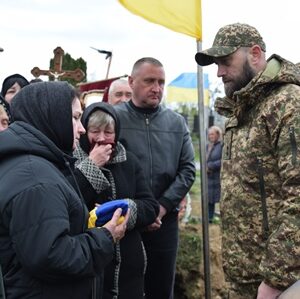 На фото зображено момент прощання: військовослужбовець у камуфляжній формі стоїть перед групою людей, серед яких жінки в темному одязі з покритими головами. Одна з жінок тримає складений синьо-жовтий прапор.