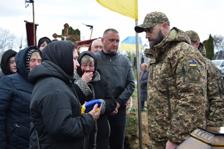 На фото зображено момент прощання: військовослужбовець у камуфляжній формі стоїть перед групою людей, серед яких жінки в темному одязі з покритими головами. Одна з жінок тримає складений синьо-жовтий прапор.