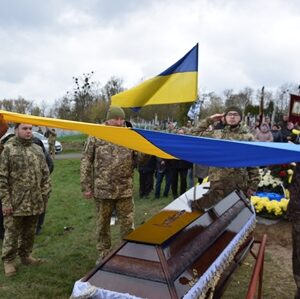 Військовослужбовці тримають державний прапор України над труною загиблого воїна під час церемонії прощання на кладовищі; поруч стоять побратими та присутні люди з квітами.