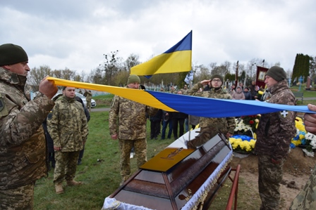 Військовослужбовці тримають державний прапор України над труною загиблого воїна під час церемонії прощання на кладовищі; поруч стоять побратими та присутні люди з квітами.