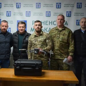 П’ятеро чоловіків, серед яких військові, стоять у приміщенні Рівненської міської ради; на столі перед ними — дрон та кейс для обладнання, поруч державні прапори.