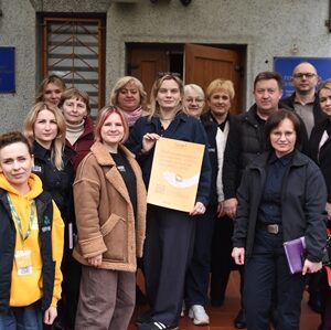 Група людей стоїть біля входу до адміністративної будівлі; у центрі жінка тримає папку з документами, учасники усміхаються та дивляться в камеру.