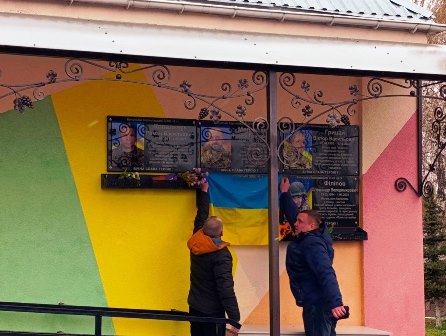 Двоє чоловіків встановлюють квіти під меморіальними дошками загиблих воїнів на стіні будівлі; поруч розміщено державний прапор України.