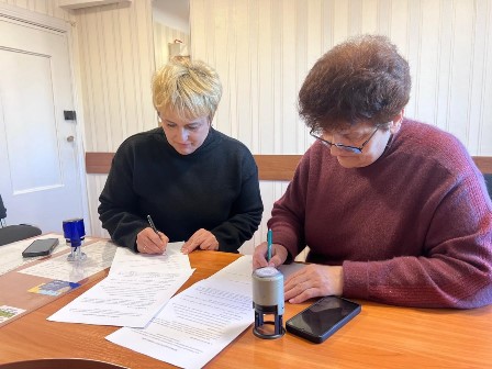 Двоє жінок сидять за столом в офісі та підписують документи. На столі лежать папери, штамп і мобільний телефон.