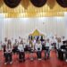 На сцені концертної зали сидить і стоїть великий учнівський музичний ансамбль. Учасники в білих сорочках і темному одязі тримають духові та ударні інструменти, перед ними встановлені пюпітри з нотами.