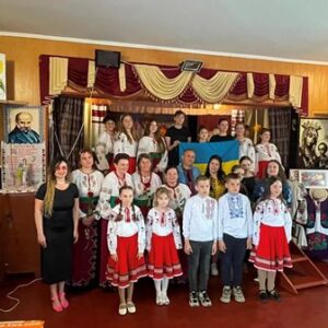 Групове фото учасників благодійного заходу «Вишийте матусю, вишиванку» у будинку культури села Карпилівка. Діти та дорослі у традиційних українських вишиванках позують на сцені з державним прапором України.
