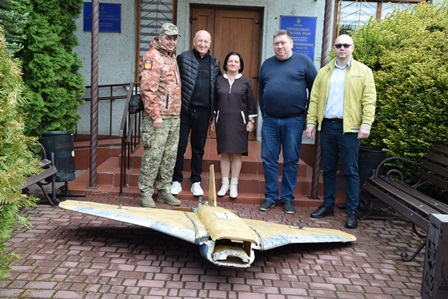 П’ятеро людей стоять біля входу до адміністративної будівлі поруч із фрагментом знешкодженого ворожого дрона «Герань», розміщеного на бруківці.