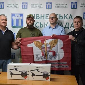 Четверо чоловіків стоять у приміщенні Рівненської міської ради, тримаючи прапор десантно-штурмових військ України. На столі перед ними розміщені два квадрокоптери в коробках.