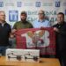 Четверо чоловіків стоять у приміщенні Рівненської міської ради, тримаючи прапор десантно-штурмових військ України. На столі перед ними розміщені два квадрокоптери в коробках.
